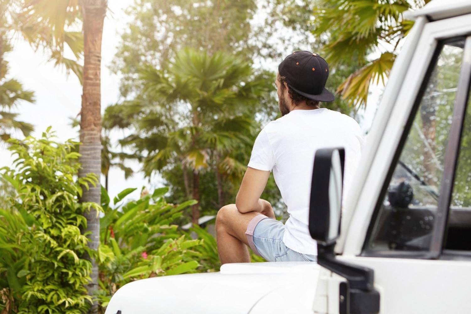 Young traveller sitting on the bonnet of a white crossover among palm trees in tropical Australia